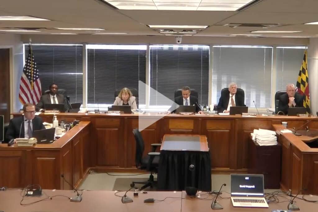 Photo of PSC Commissioners during a Proceeding, with a large "play" arrow superimposed on top of the image, signifying that clicking the image will lead to a video.